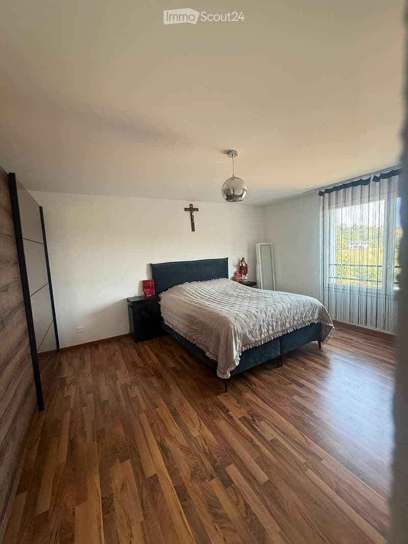 Spacious bedroom with wooden flooring, large bed with dark headboard and white bedding, nightstand with candle, mirror, and decorative items, large window with sheer curtains, and a cross on the wall.