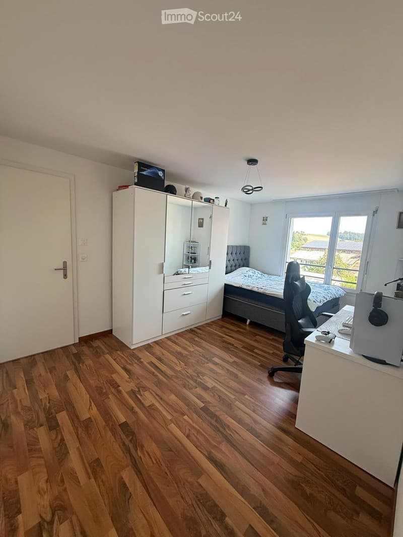 A well-lit bedroom with wooden flooring, a large bed with a patterned duvet, a wardrobe with a mirror, a desk with a computer, and a chair.
