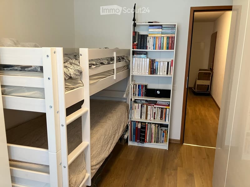 Cozy bedroom with a white bunk bed and a bookshelf filled with books. The lower bunk has a beige blanket, and the room features wooden flooring.