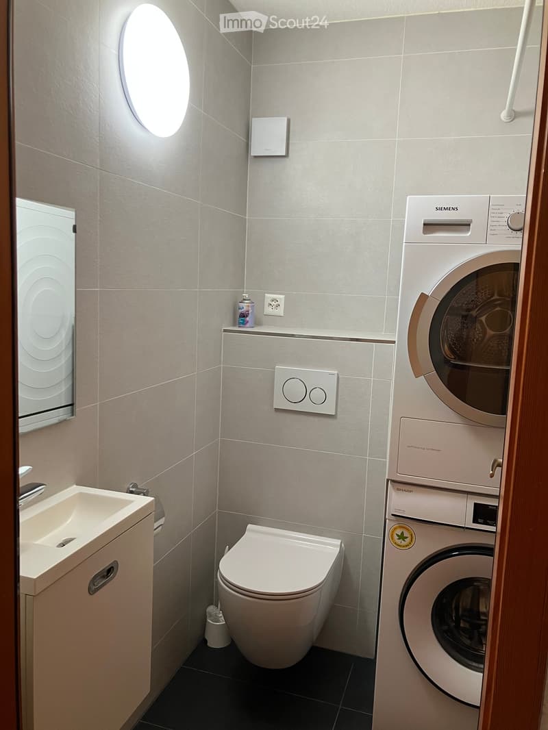 Modern bathroom with a white sink, toilet, washing machine, and a round window. The walls are tiled in light gray, and the floor is dark gray.