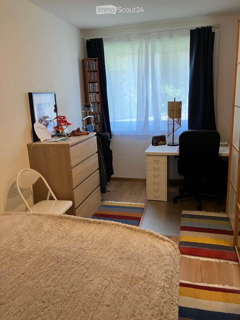 Cozy bedroom with a fluffy bed, a wooden desk, a chair, a bookcase, and a small table lamp. The room features a large window with dark curtains and a colorful striped rug.