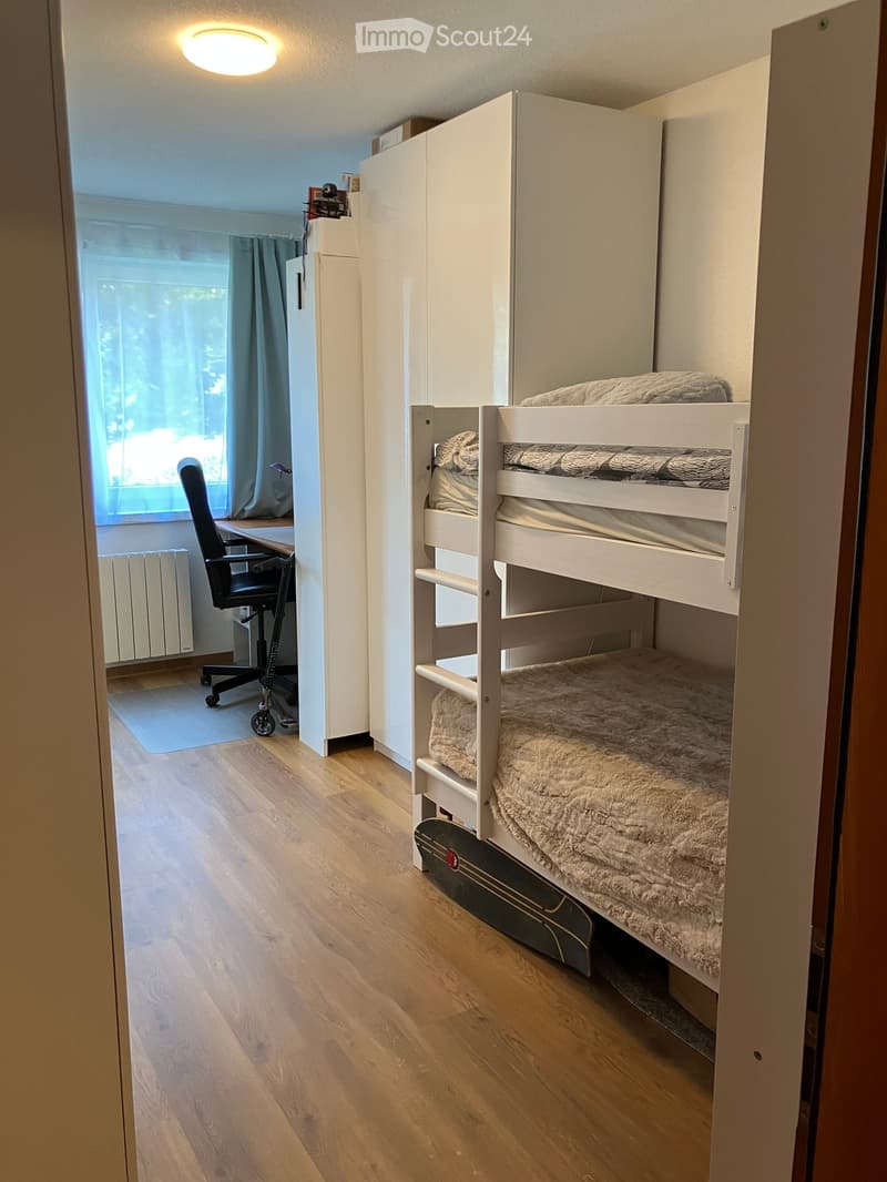 Cozy bedroom with a white bunk bed, a desk with a chair, and a wardrobe. The room has wooden flooring and large windows with blue curtains.