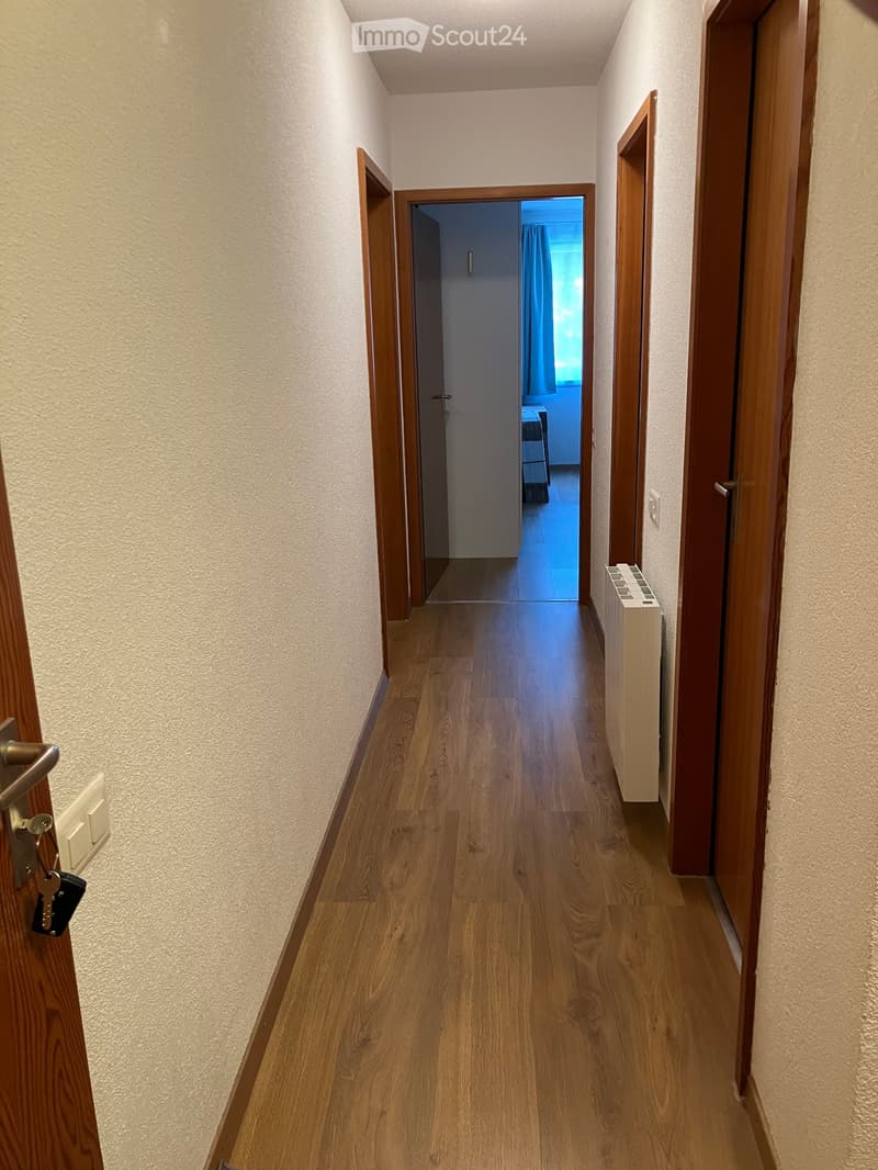 A narrow hallway with wooden flooring and light-colored walls, featuring several doors and a radiator on one side.