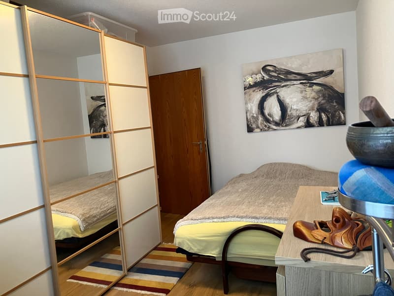 A small, cozy bedroom with a bed, a small table, a stool, and a painting on the wall. The room features a sliding door and a colorful rug on the floor.
