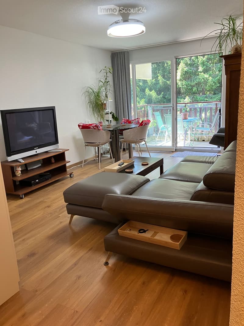 Modern living room with hardwood floors, a grey leather sofa, coffee table, TV, and a dining area with chairs and a table. Large windows and a sliding glass door lead to a balcony with outdoor furniture.