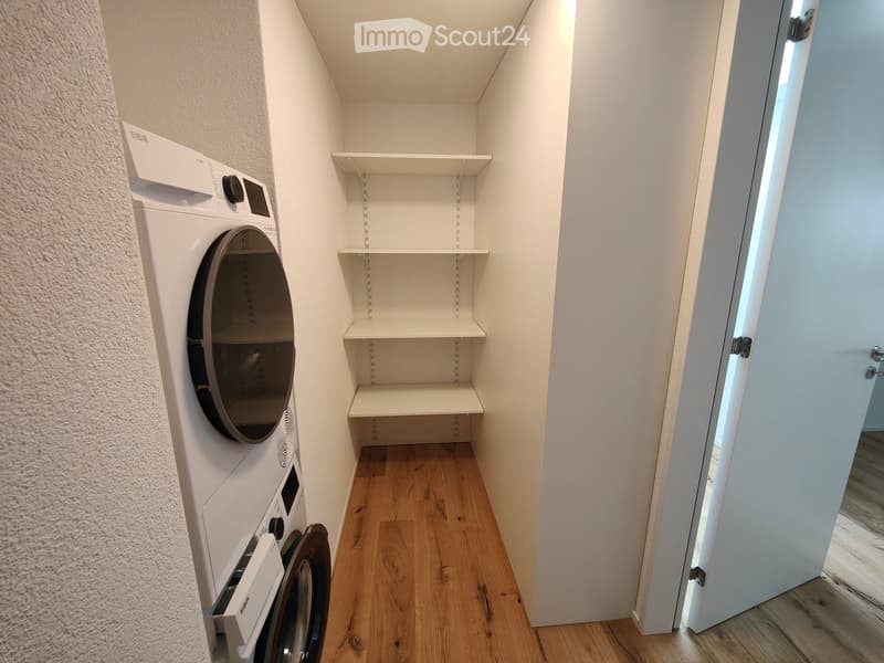 Empty room with wooden flooring, white walls, built-in shelving, and a private washing machine installed in the corner.