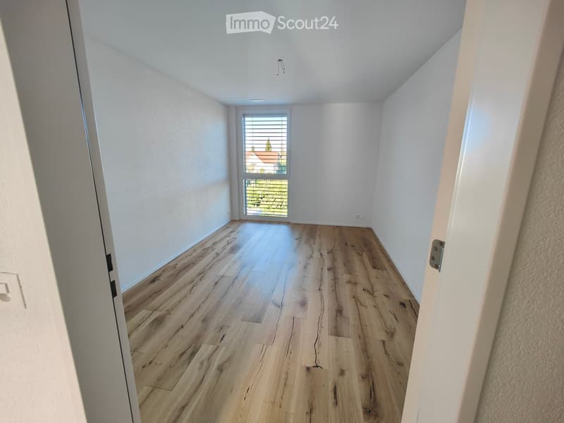 Empty room with light wooden flooring and white walls, featuring a window with blinds and a visible door frame.