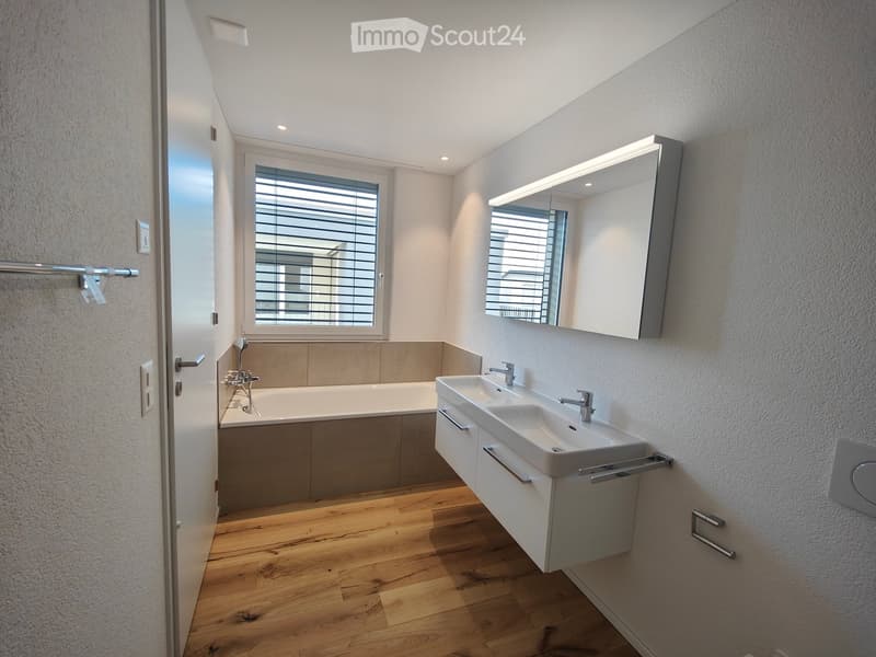 Modern bathroom with a bathtub, double sink, large mirror, and wooden flooring.