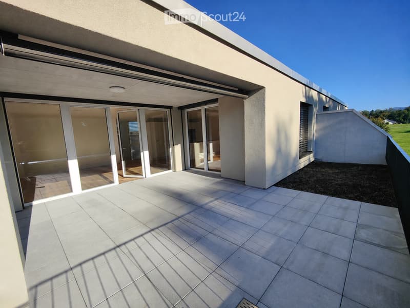Modern balcony with large sliding glass doors leading to an interior space, tiled floor, and a view of a green field and mountains in the background.