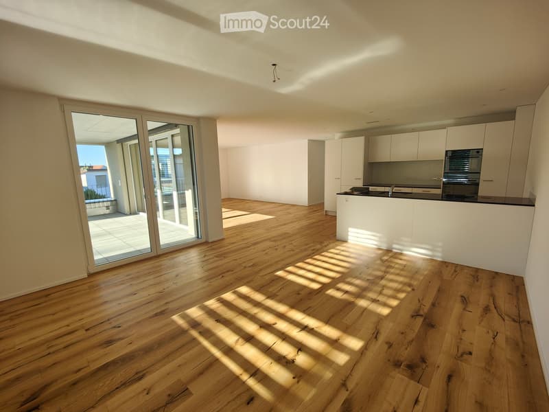 Open-plan unfurnished room with light wooden flooring, white walls, and a modern kitchen area featuring white cabinets and stainless steel appliances. Large sliding glass doors lead to a balcony.