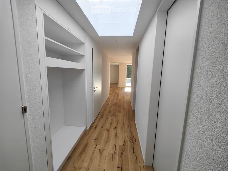 Modern hallway with wooden flooring, white walls, built-in shelving on one side, and a skylight at the far end.