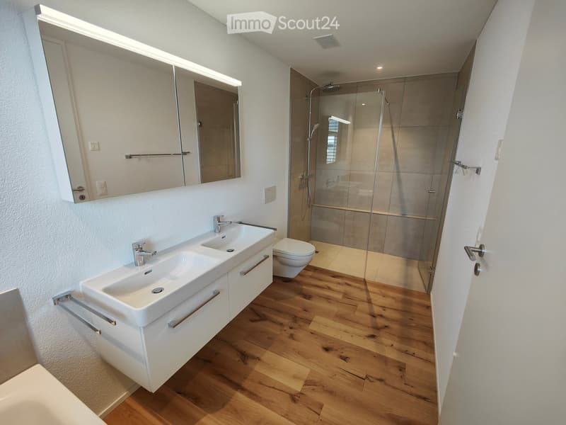 Modern bathroom with double sink, mirror, toilet, and glass-enclosed shower. Wooden flooring and white cabinetry.