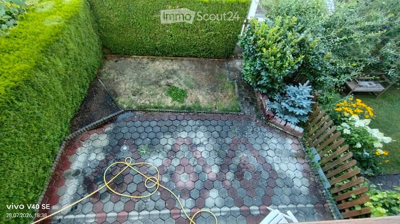 Garden area with a paved patio, surrounded by hedges and greenery, featuring a small wooden bench and a coiled yellow hose.
