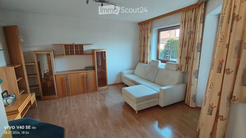 Living room with light-colored sofa, ottoman, wooden cabinet, shelves, and a mirror. The room has wooden flooring and large windows with curtains.