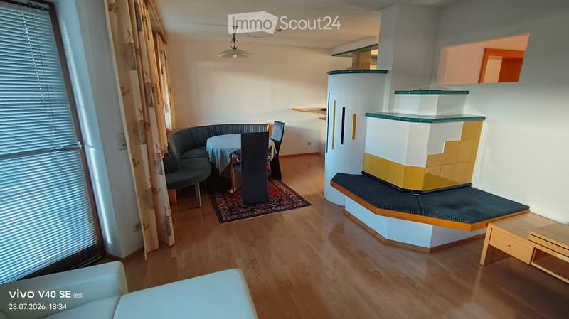A modern living room with a sofa, dining table, chairs, fireplace, and small table. The room features wooden flooring and large windows with curtains.