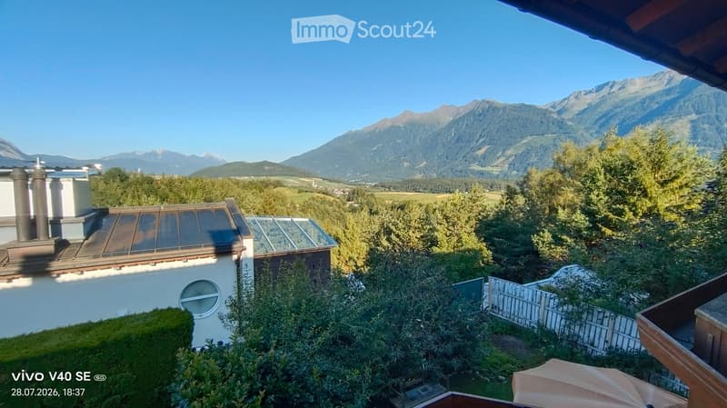 A scenic outdoor view from a balcony or terrace, showing a lush garden area with trees and shrubs, a white fence, and a distant mountain range under a clear blue sky.