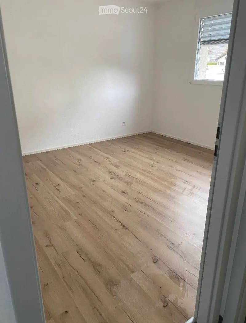 Empty room with light wooden flooring and white walls, featuring a window with blinds and a visible electrical outlet.