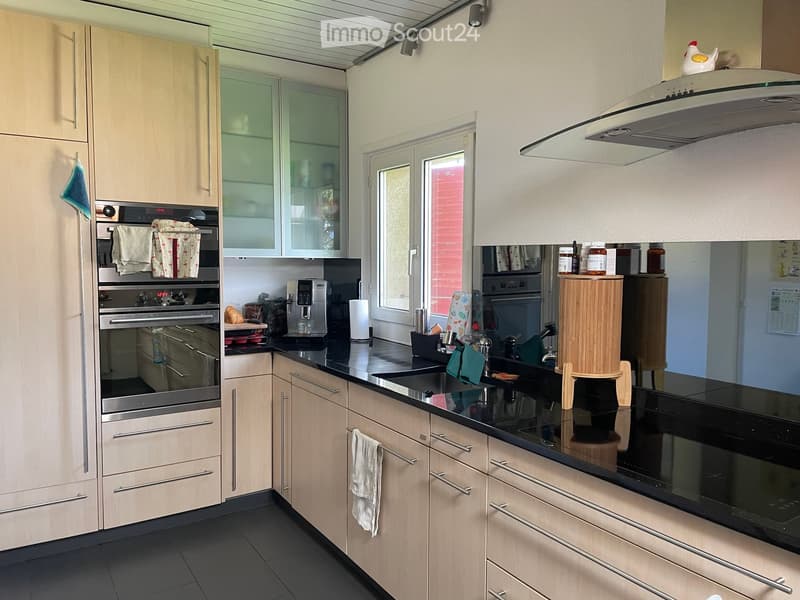 Modern kitchen with light wood cabinets, stainless steel appliances, black countertops, and a window overlooking the outside.
