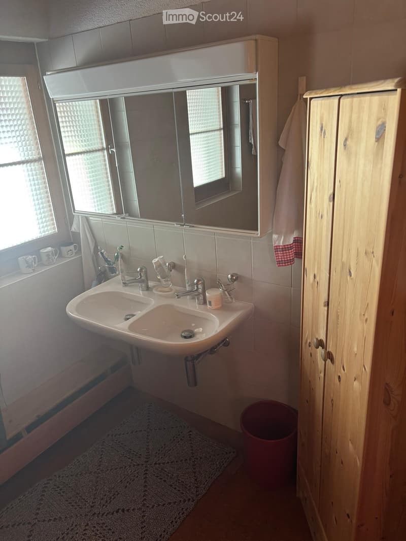 Modern bathroom with a double sink, mirror cabinet, wooden cupboard, and a small window with frosted glass. The sink has two faucets and various toiletries, including toothbrushes and toothpaste. A towel hangs on the cupboard, and a small rug is placed on