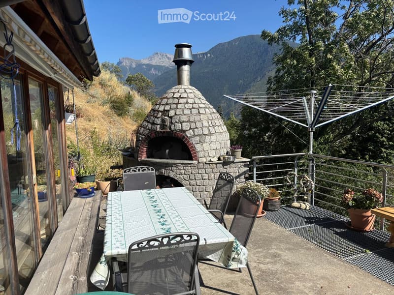 Balcony with a stone oven, a table with chairs, and potted plants, overlooking a scenic mountain view.