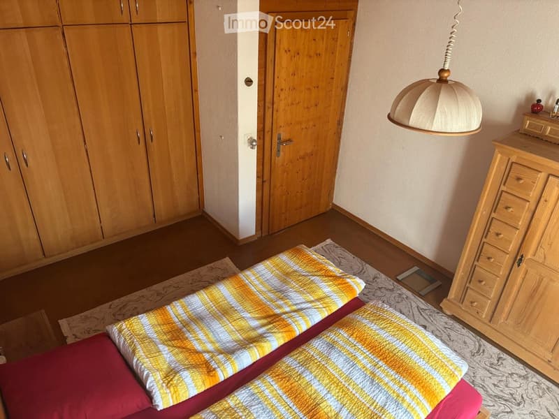 Bedroom with a neatly made bed, wooden wardrobe, dresser, and a hanging lamp.