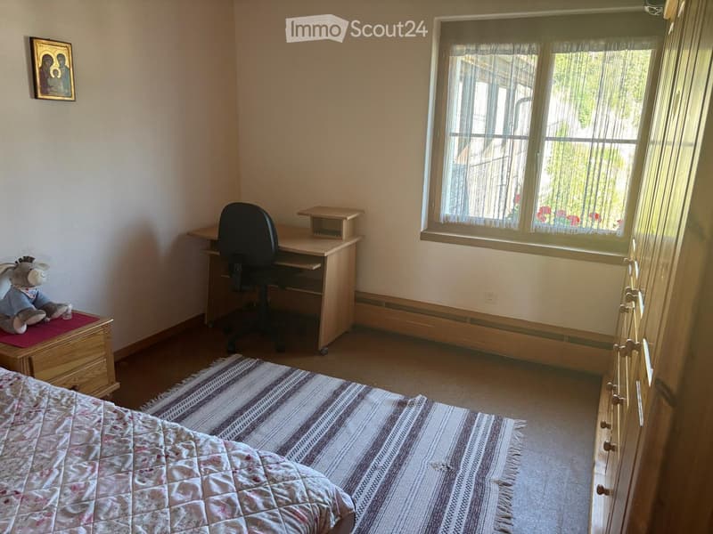 Cozy bedroom with a bed, desk, chair, and a small wooden cabinet. A colorful rug lies on the floor, and a framed picture hangs on the wall. A stuffed animal sits on the cabinet.