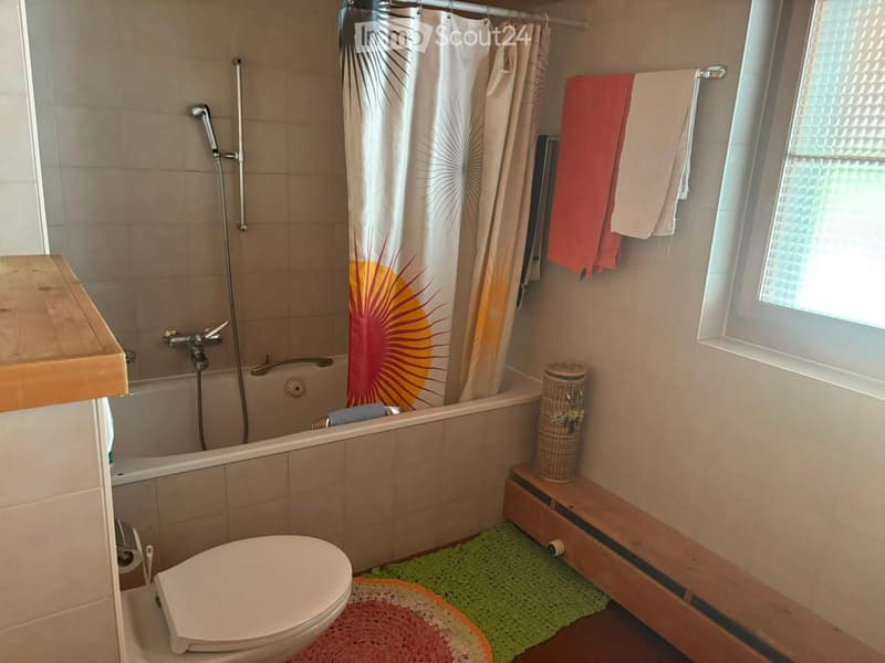 Bathroom with a toilet, bathtub, shower curtain, towels, a wooden bench, a basket, and rugs.