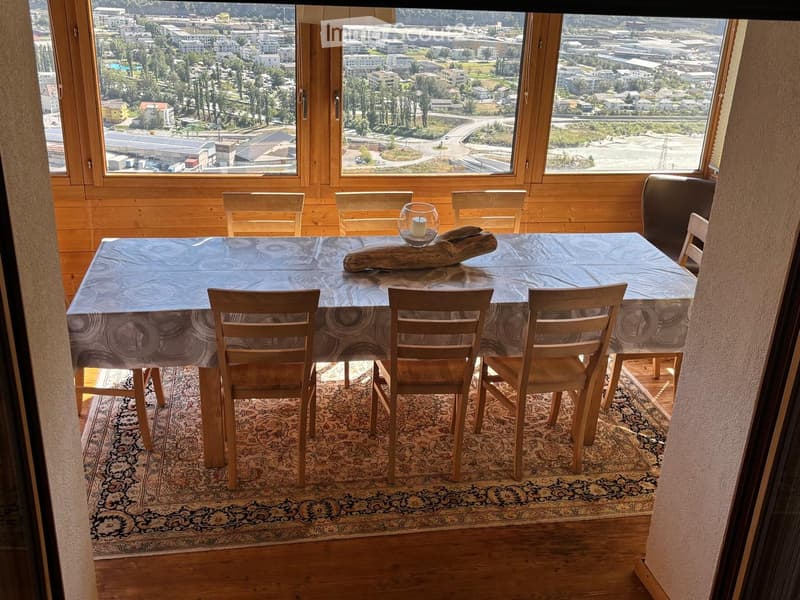 Dining area with a wooden table, six chairs, and a decorative candle, overlooking a cityscape.
