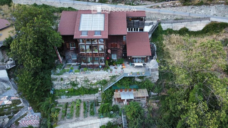 A multi-story wooden house with red-tiled roofs, large windows, and a stone terrace overlooking a hillside. The house is surrounded by greenery and has a small garden area below.