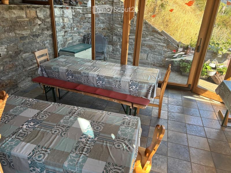 A cozy dining area with a table covered in a patterned tablecloth, surrounded by wooden chairs and a bench, with large windows offering a view of the outdoor area.