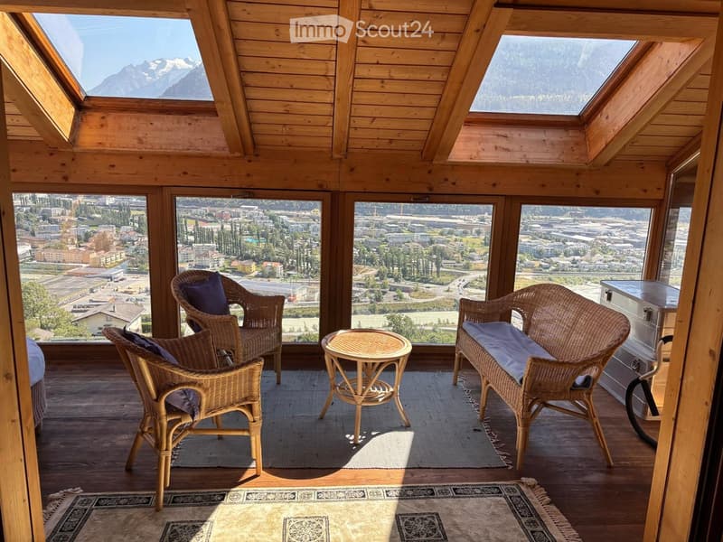 Wooden balcony with panoramic city and mountain views, featuring wicker chairs, a small round table, and patterned rugs.