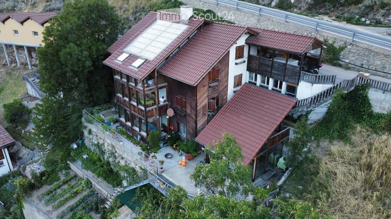 A multi-story residential building with red-tiled roofs, wooden balconies, and a garden area. The structure includes multiple levels with large windows and outdoor spaces.