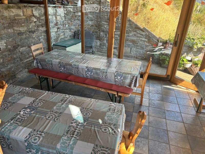 A cozy dining area with a table covered in a patterned tablecloth, surrounded by wooden chairs and a bench, with large windows offering a view of the outdoor area.