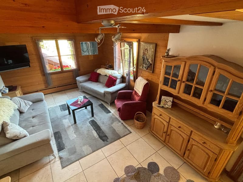 Cozy living room with wooden walls and ceiling, featuring a grey sectional sofa, red armchair, coffee table, wooden cabinet, and large windows with curtains.