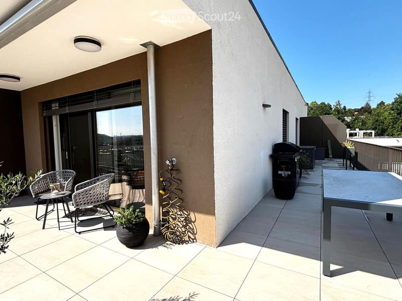 Modern balcony with tiled floor, white walls, and a view of greenery. Features two wicker chairs, a small table, potted plants, and a grill.