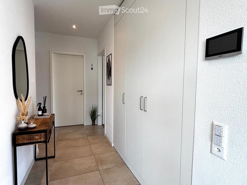 Modern hallway with tiled floor, white walls, built-in closet, small table with decorative items, and a wall-mounted TV.