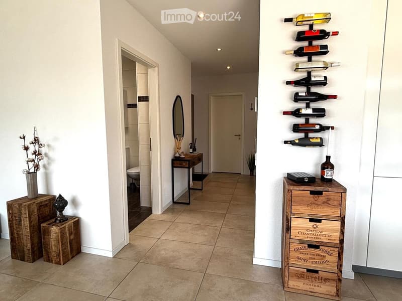 Modern hallway with tiled floor, white walls, recessed lighting, and a wine rack on the right side. A small table with a vase of flowers and a mirror is visible, along with a bathroom entrance.