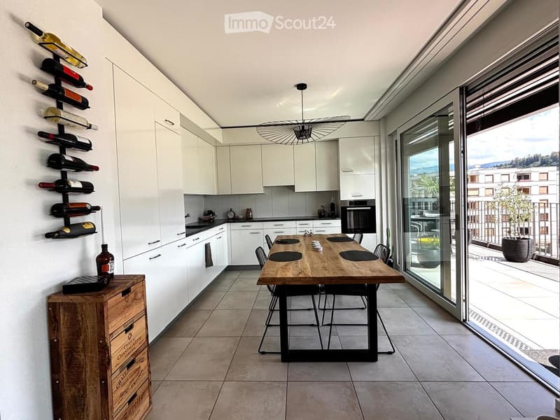 Modern kitchen with white cabinets, a wooden dining table, black chairs, and a wine rack on the wall. The space features large windows with a view of the balcony and outdoor area.