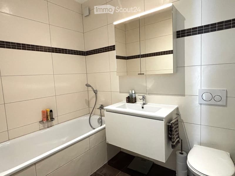 Modern bathroom with a bathtub, sink, mirror, and toilet. Beige tiles with black accents, a built-in sink cabinet, and a towel hanging on the side.