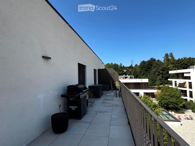 Modern balcony with tiled floor, barbecue, potted plant, and railing overlooking a landscaped area with trees and modern buildings.