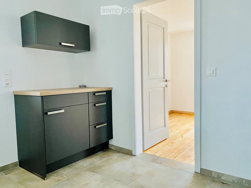 Modern kitchen with grey cabinets, a wooden countertop, and a tiled floor. An open doorway leads to another room with wooden flooring.