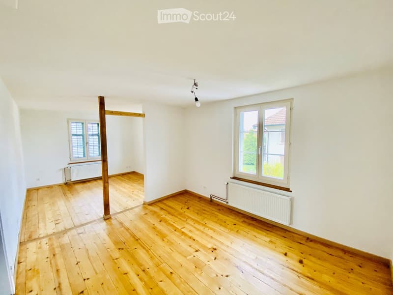 Empty room with wooden flooring, white walls, and two windows with radiators underneath. A wooden beam is visible in the center.