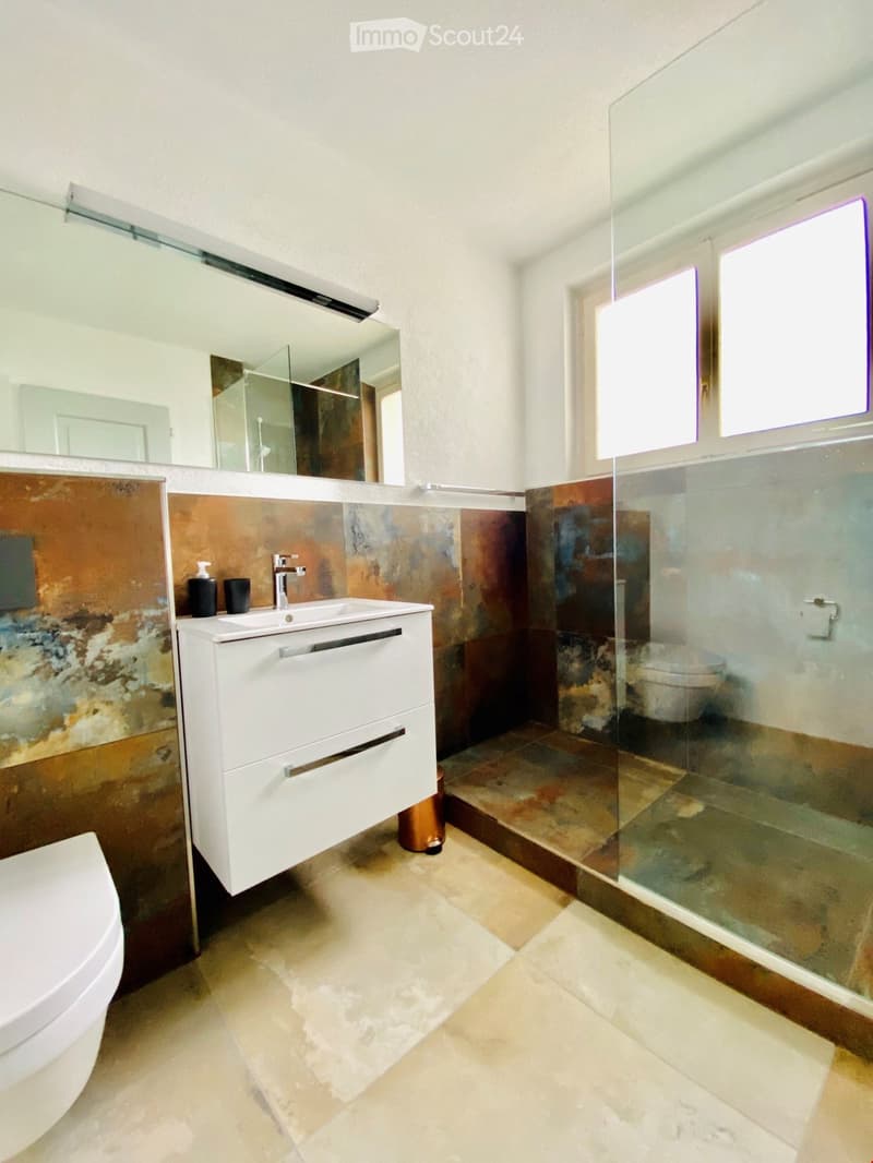 Modern bathroom with a white sink cabinet, a glass-enclosed shower, and a toilet. The walls feature a mix of brown and beige tiles, and the floor is tiled in a light beige color.