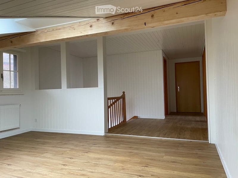 Empty room with wooden flooring, white paneled walls, and a visible staircase leading to another level. The room has a window on one side and a radiator below it.