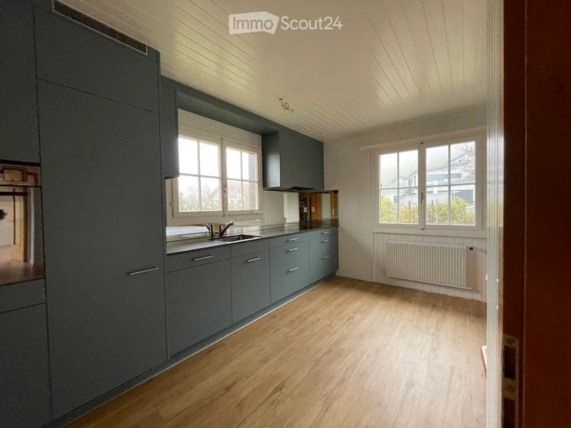 Empty kitchen with grey cabinets, wooden flooring, and large windows. No appliances or furniture are present.