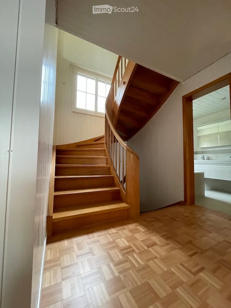 Spacious hallway with wooden flooring and a wooden staircase leading to an upper level. A window provides natural light, and a partially visible bathroom is seen to the right.