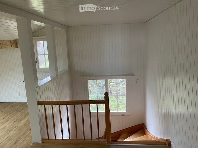 Spacious, well-lit hallway with wooden flooring and white paneled walls. Features a wooden staircase with a simple railing leading upwards, and a window providing natural light.
