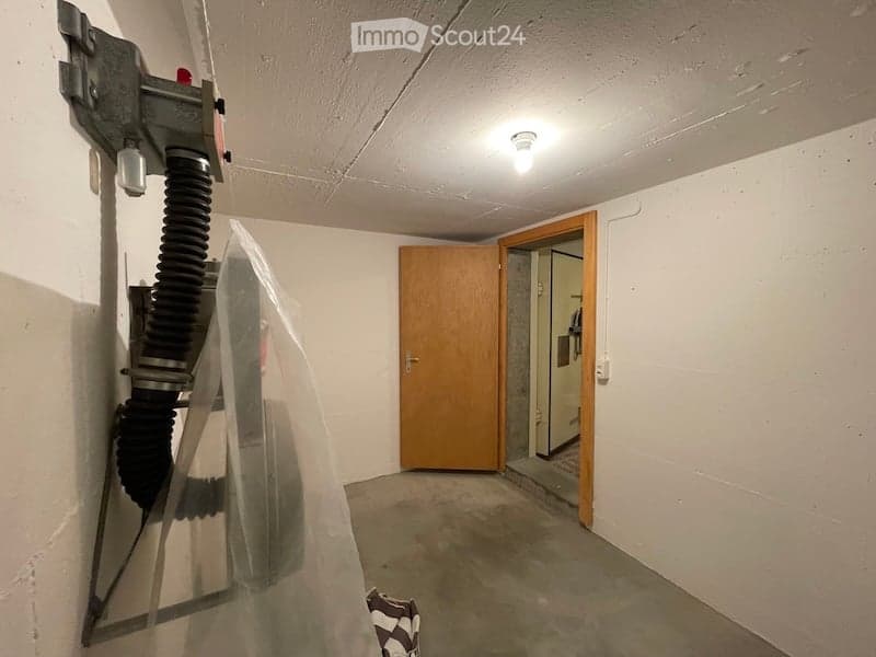 Empty room with white walls, concrete floor, and a wooden door leading to another space. A vacuum cleaner is partially visible on the left side.