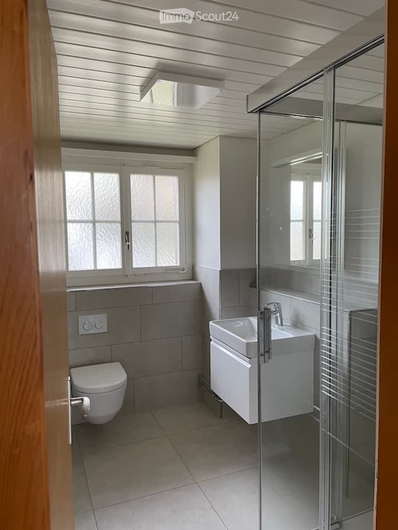 Modern bathroom with white tiled floor, grey tiled walls, a toilet, a sink with a chrome faucet, and a glass-enclosed shower. The room features a window with frosted glass panes and a sleek, contemporary design.