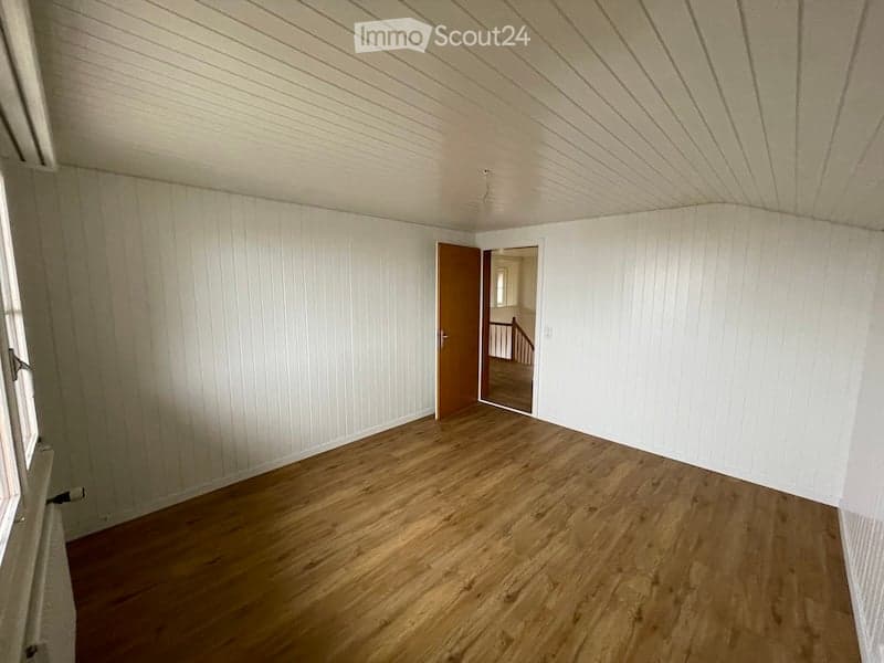 Empty room with wooden flooring, white paneled walls, and a door leading to another room. No furniture or equipment is present.